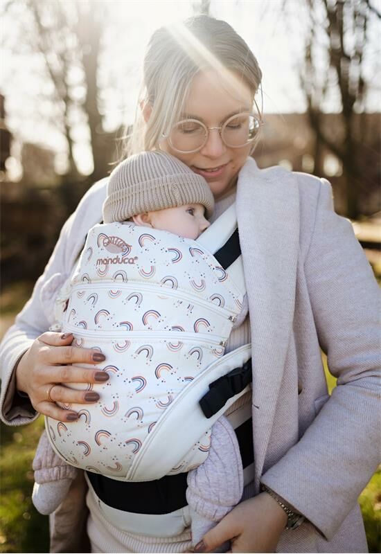 A woman with blond hair, gold glasses, wearing a grey coat, standing outside carrying her baby in a Manduca XT Limited Edition Rainbow Day Baby Carrier.