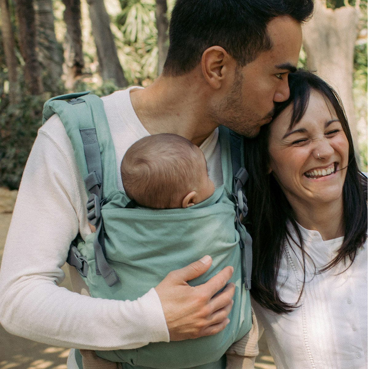 A couple hugging and laughing together whilst the guy carries their newborn in a Boba X Baby Carrier.