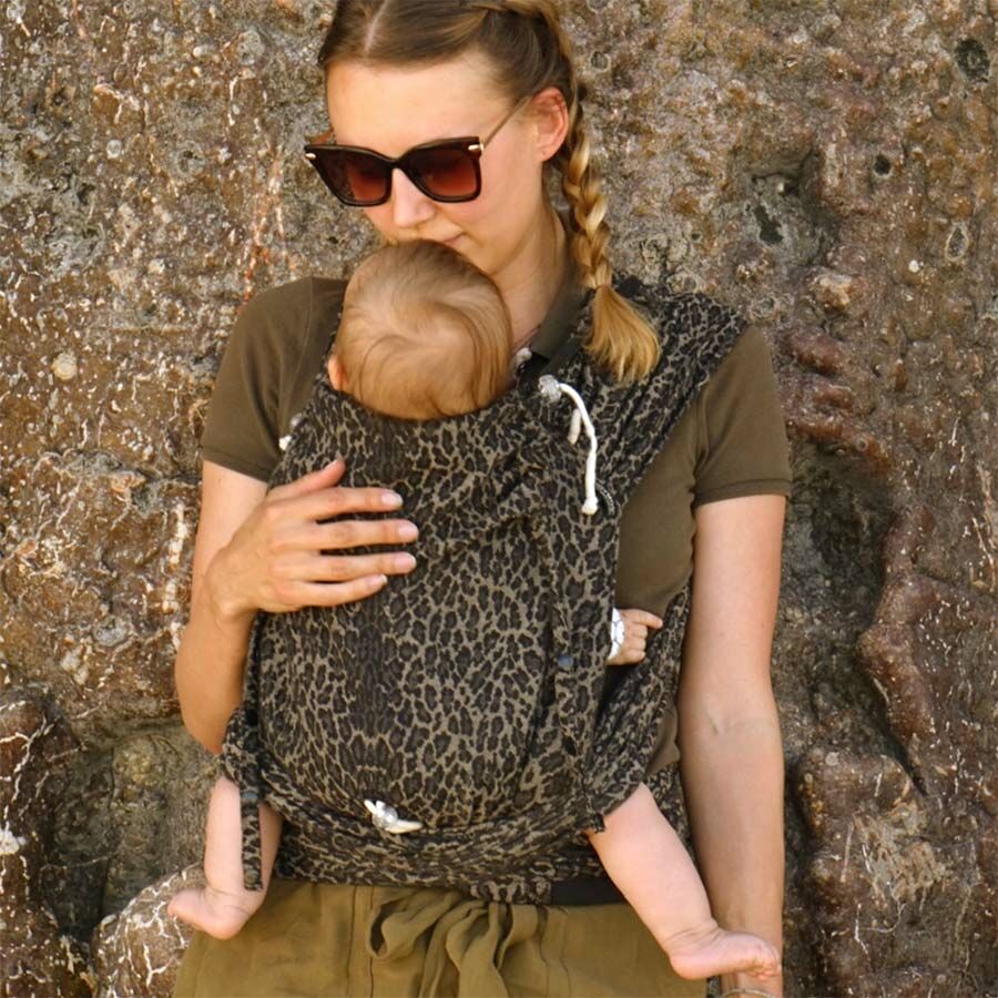 Woman with her hair in pleats wearing sun glasses standing in front of a stone wall carrying her baby in a Didymos Didyklick Leo Half Buckle Carrier.