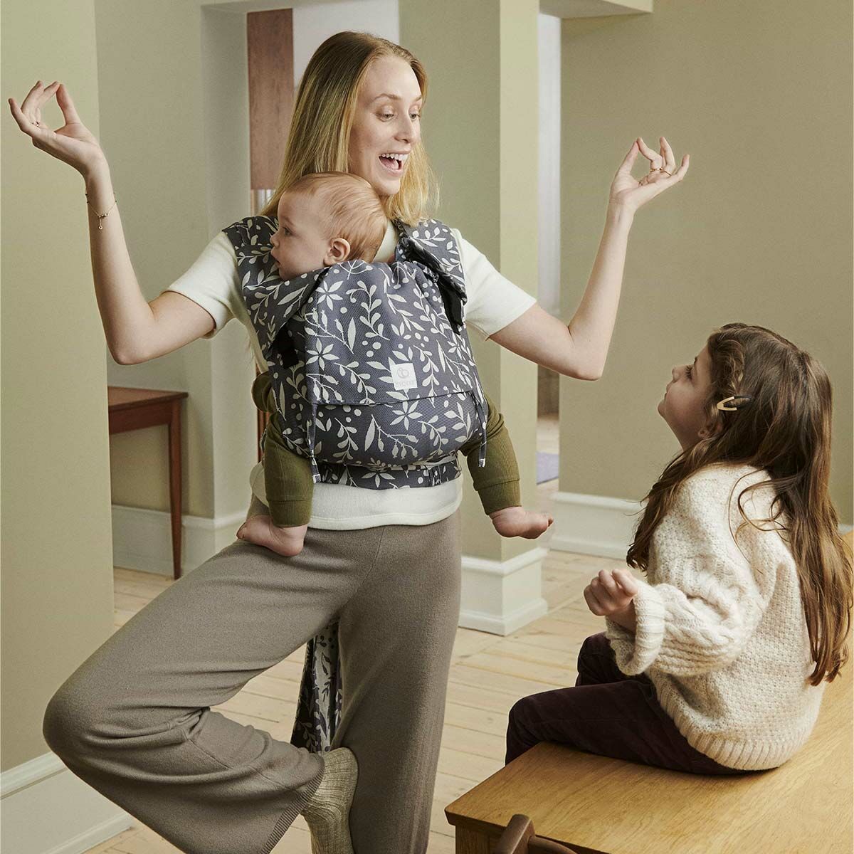 A woman standing in a living room carrying her baby in a Stokke Limas baby carrier whilst entertaining her young daughter who is sitting in front of her.