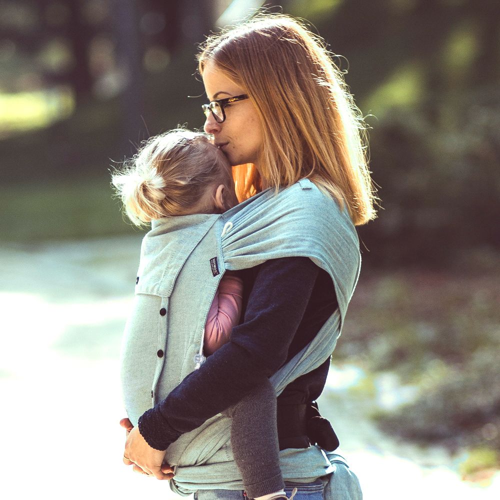 A woman with brown hair and glasses wearing a navy top and blue jeans standing outside on a sunny day carrying her toddler daughter on her front in a Didymos Didyklick 
