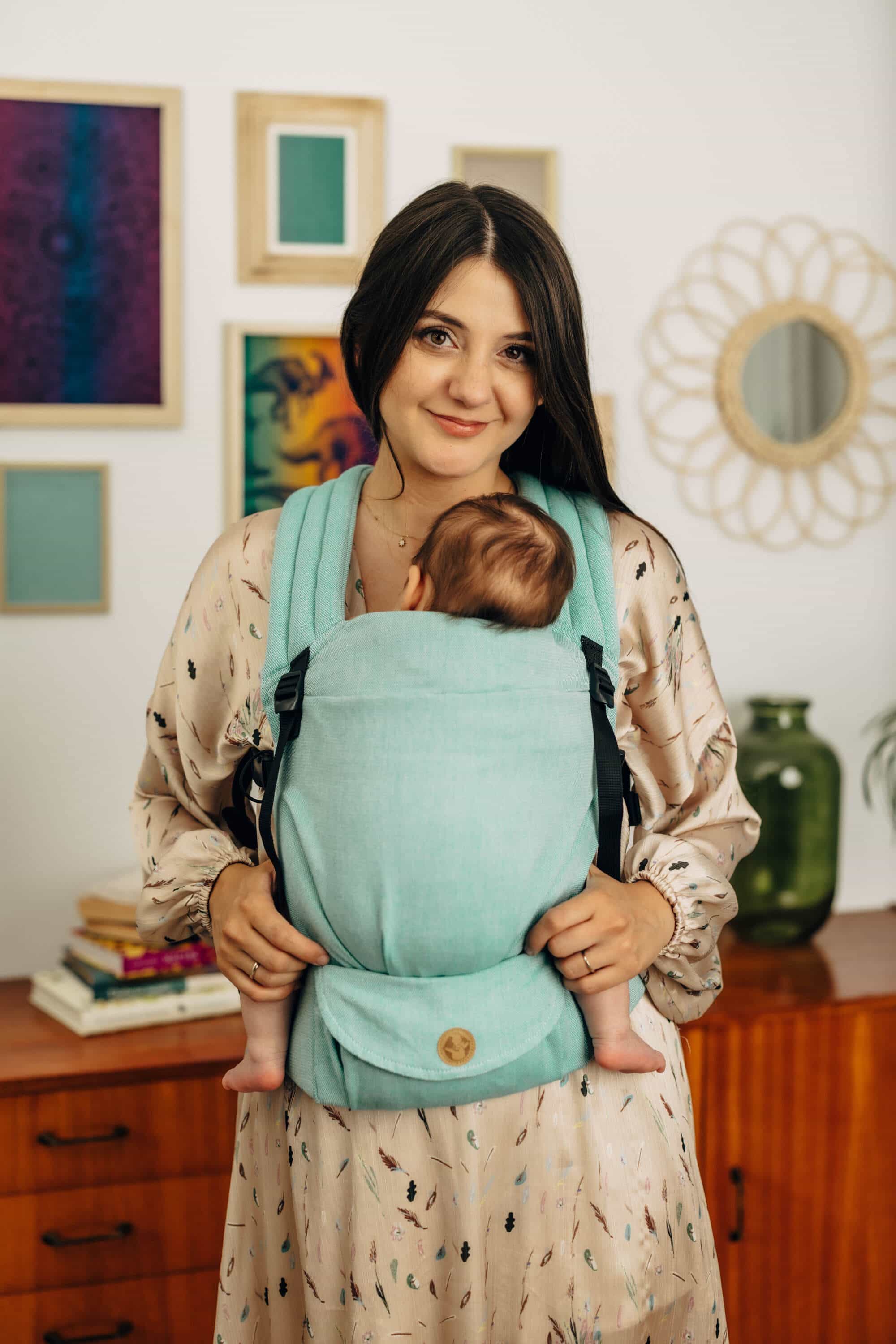 A woman with brown hair carrying her young baby in a Lennylight Agave baby carrier.