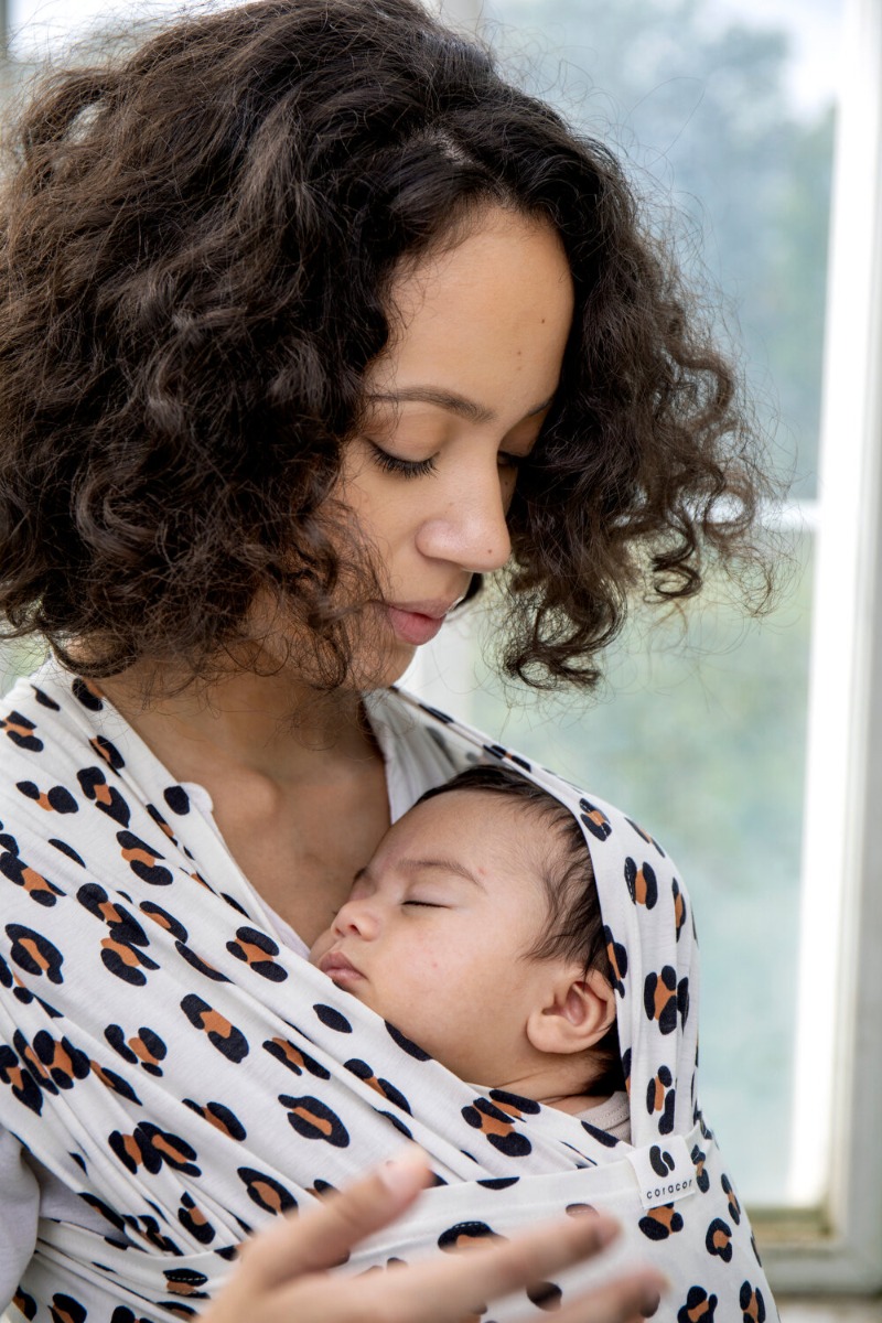 A close-up image of a woman with dark, curly hair, looking down at her newborn baby whilst carrying her in a white baby wrap sling with a leopard print.