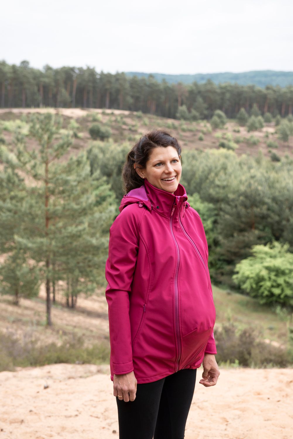 A woman with dark hair in a ponytail standing outdoors on a hill wearing the Mamalila Allrounder Babwywearing Jacket in Berry with the maternity insert zipped in.