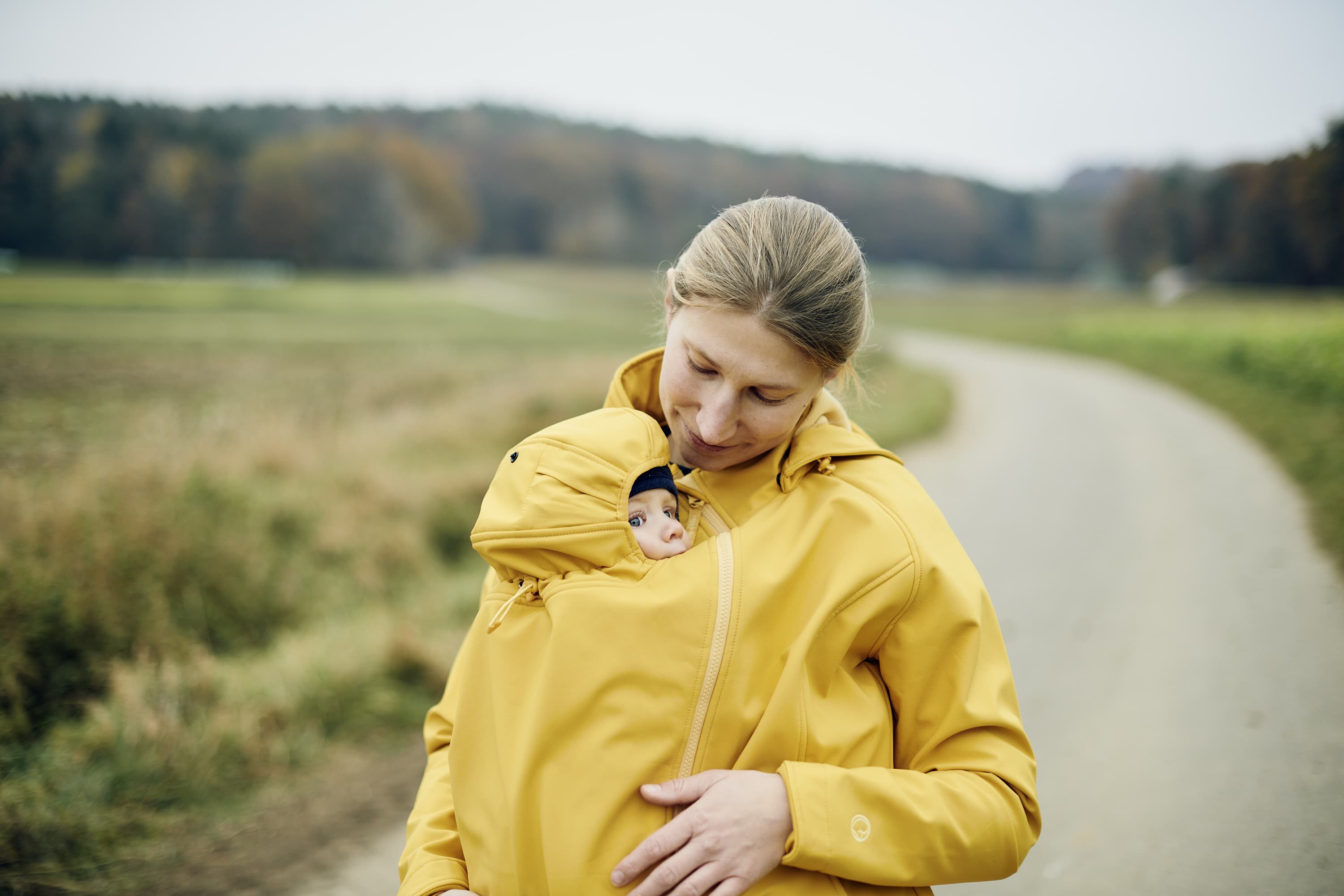 A woman with blonde hair tied back in a pony tail standing outside looking down lovingly at her baby wearing the Mamalila Allrounder Babywearing Jacket in Mustard with her baby in the babywearing insert.