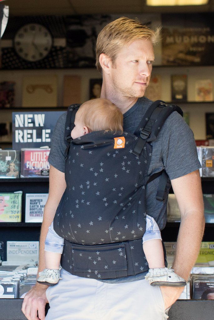 Best Toddler Carriers 2023. A man wearing a grey t-shirt and white trousers, looking to his left carrying his baby daughter in a Tula Toddler Carrier Discover.