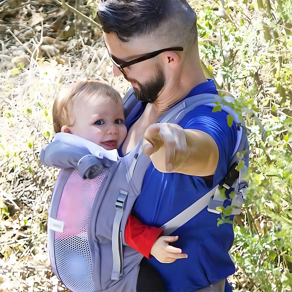 A man with dark hair and a beard wearing sunglasses and a blue t-shirt standing in a sunny forest and pointing at the camera, carrying his blond-haired son in a grey Love Radius Physiocarrier with the panel rolled away and using the mesh panel.
