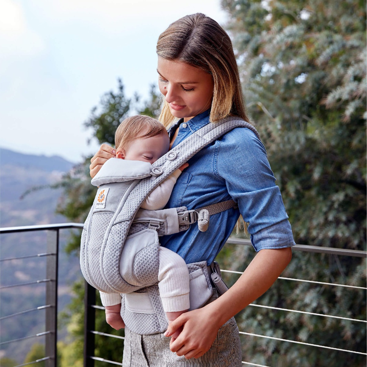 Babywearing in Summer - 8 Top Tips For Staying Safe & Cool. A woman with long dark hair, wearing a blue denim shirt and grey striped trousers, stands on a balcony in front of lush green vegetation on a sunny day, and stares adoringly at her sleeping, blond-haired baby son whom she is carrying on her front in a Grey Ergobaby Omni Breeze Baby Carrier.