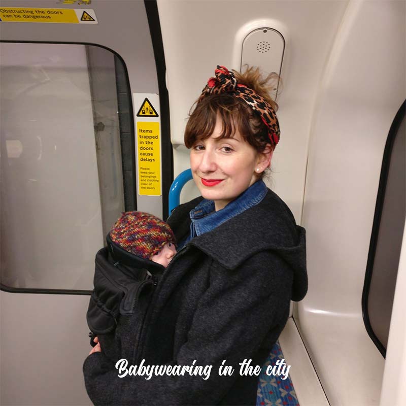 Katie sitting on a subway train with her baby in a sling and wearing a Mamalila Wool Hooded Coat