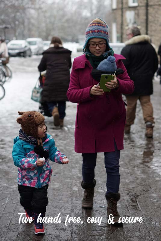 Dee walking along a snowy street carrying her baby in a sling wearing a Mamalila Wool Hooded coat and her toddler walking along beside her.