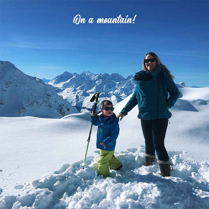 Rachel on top of a snowy mountain in Switzerland on a sunny day with her baby in a sling and wearing a Blue Mamalila Babywearing Softshell Jacket Allrounder and her young son standing in the snow beside her.