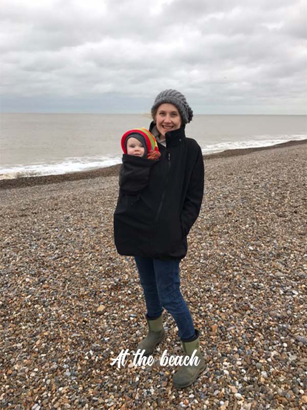 Helen standing on a stony beach carrying her baby in a sling and wearing a black Mamalila Babywearing Softshell Jacket Allrounder