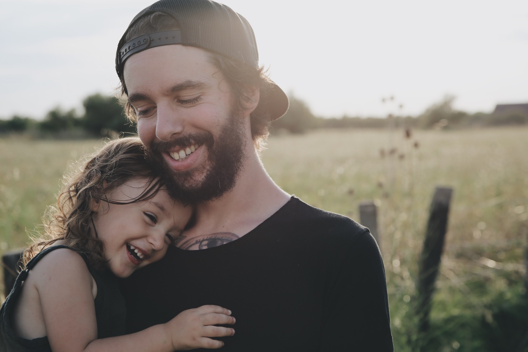A close-up image of a bearded, dark-haired man wearing a cap backwards and a black t-shirt, cuddling and laughing with his young daughter outdoors in a field on a sunny day.
