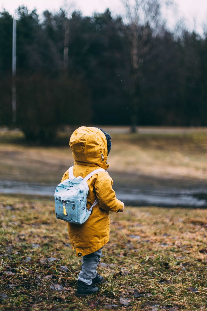A small child wearing a yellow jacket with the hood up and a blue back pack walking away from the camera