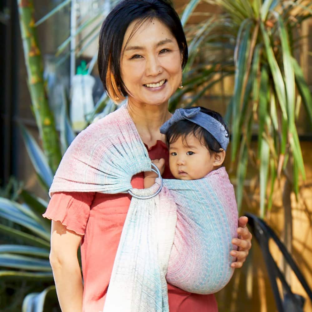 A smiling, dark-haired Japanese woman wearing a pink t-shirt, standing outside in the sunshine in front of palm trees, carrying her dark-haired baby daughter on her left hip in a Didymos Prima Aurora Didysling