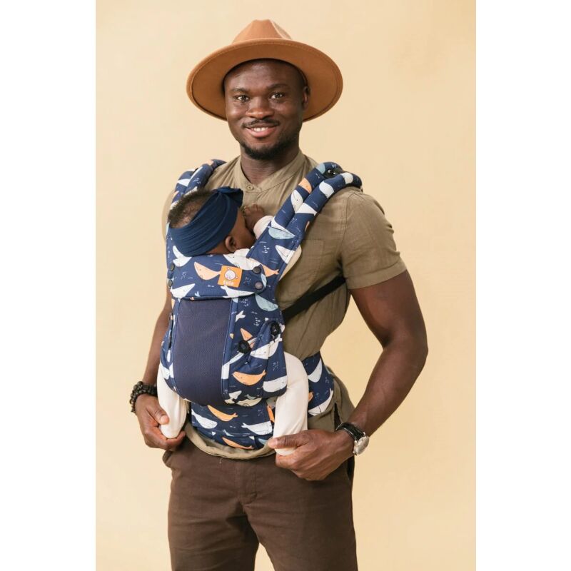 A smiling, bearded black man wearing a khaki shirt, brown jeans and a khaki hat, carrying his baby daughter in a Tula Coast Explore Whale Watch baby carrier.