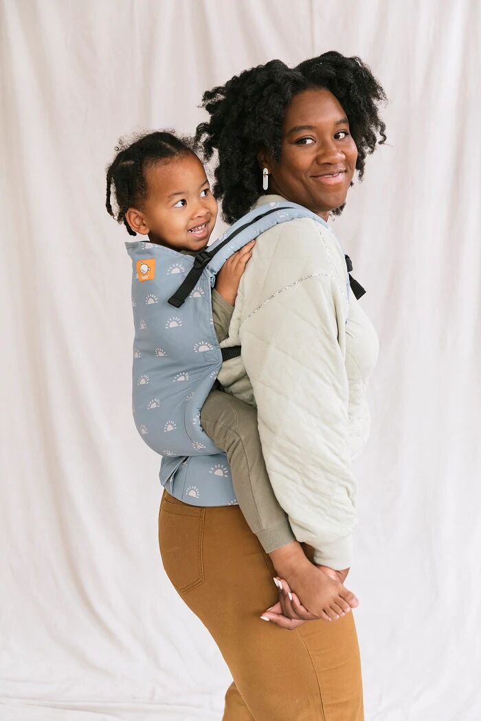 A woman with black hair wearing a cream top and brown trousers carrying her toddler in a Tula Harbor Skies Toddler Carrier