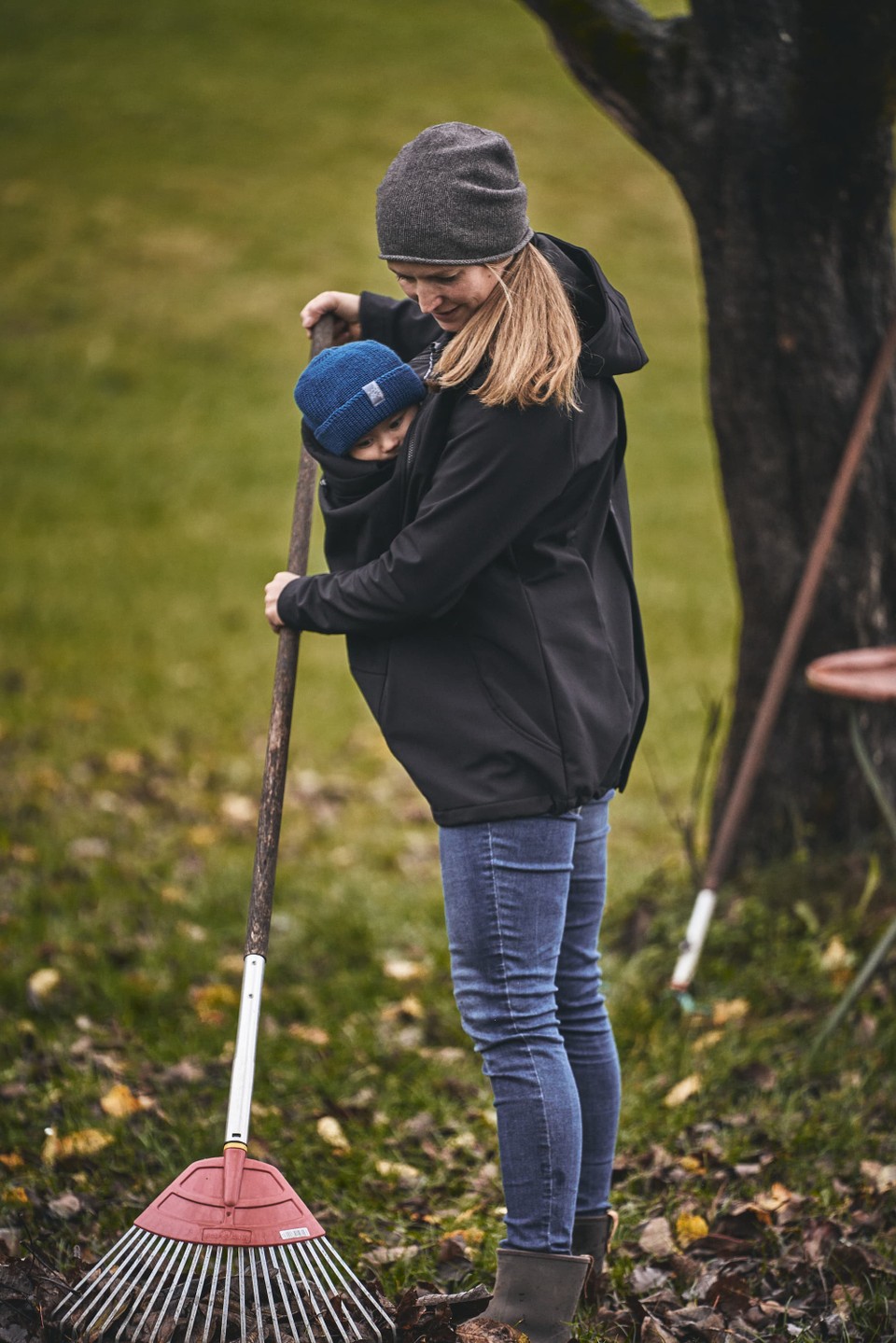 A woman with blonde hair wearing a grey beanie hat with her baby in a Mamalila Allrounder Babywearing jacket with the babywearing insert carrying her baby in a sling who is wearing a blue hat, outside in the garden raking leaves.