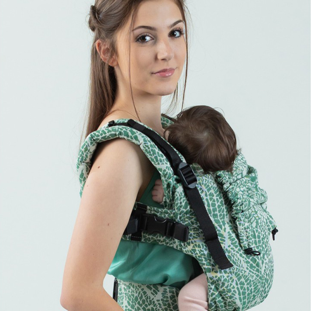 A photo of a young mum carrying her baby in an Isara The One baby carrier, indoors with a grey background.