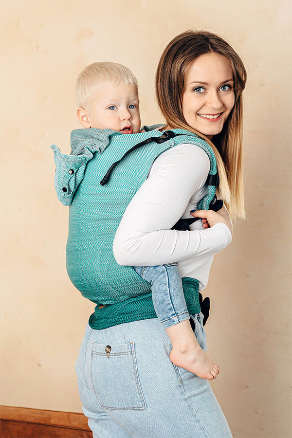 A woman with long brown hair wearing a white top and blue jeans carrying her toddler on her back in a LennyLamb LennyGo Ombre Green carrier.