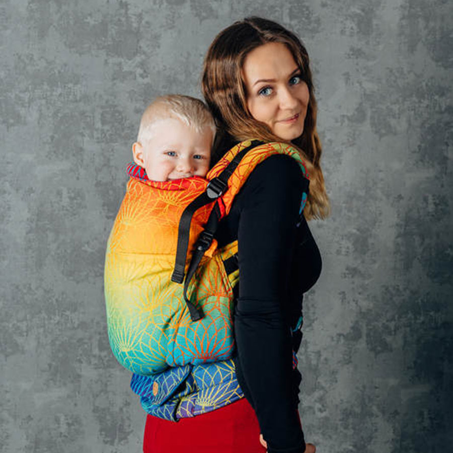 A woman with long brown hair wearing a black top and red trousers, standing indoors in front of a grey mural, looking over her right shoulder smiling at the camera as she carries her blond-haired toddler son on her back in a Lenny Lamb Rainbow Lotus Preschool Carrier 