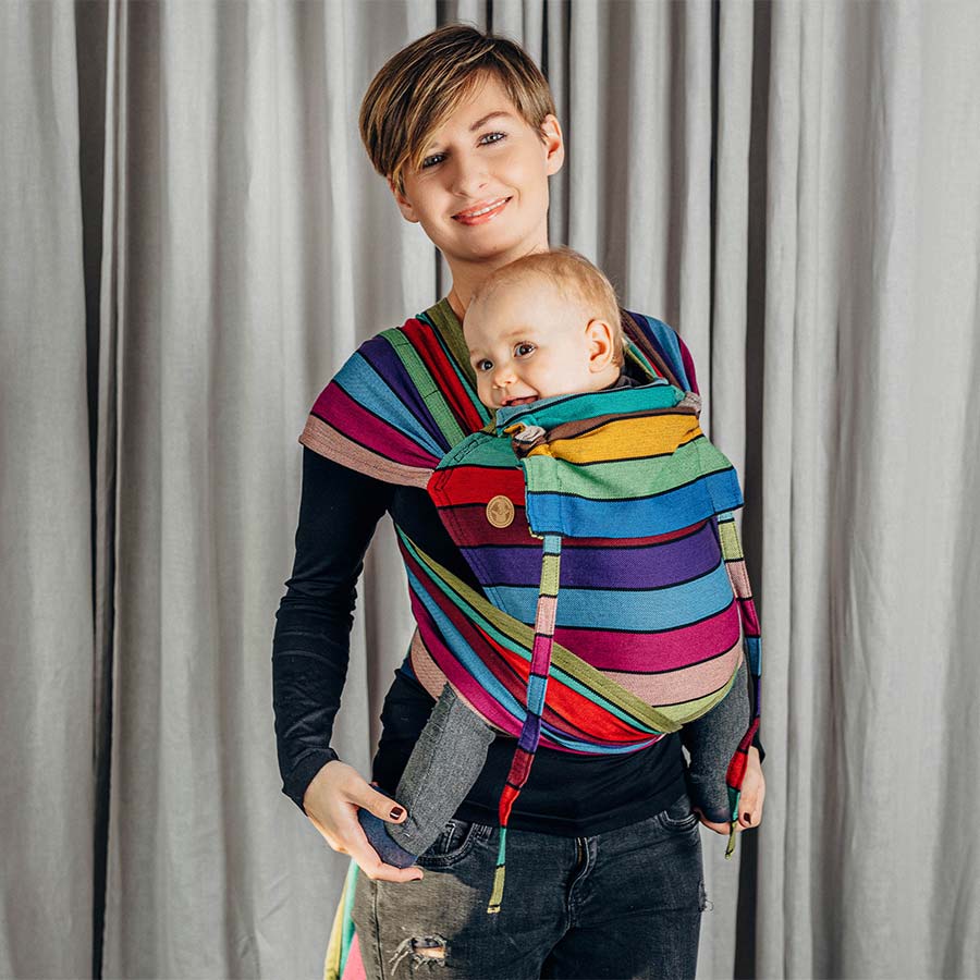 A woman with short hair wearing a black top and black jeans, standing indoors facing the camera carrying her toddler son in a Lenny lamb Wraptai Carousel of Colours.
