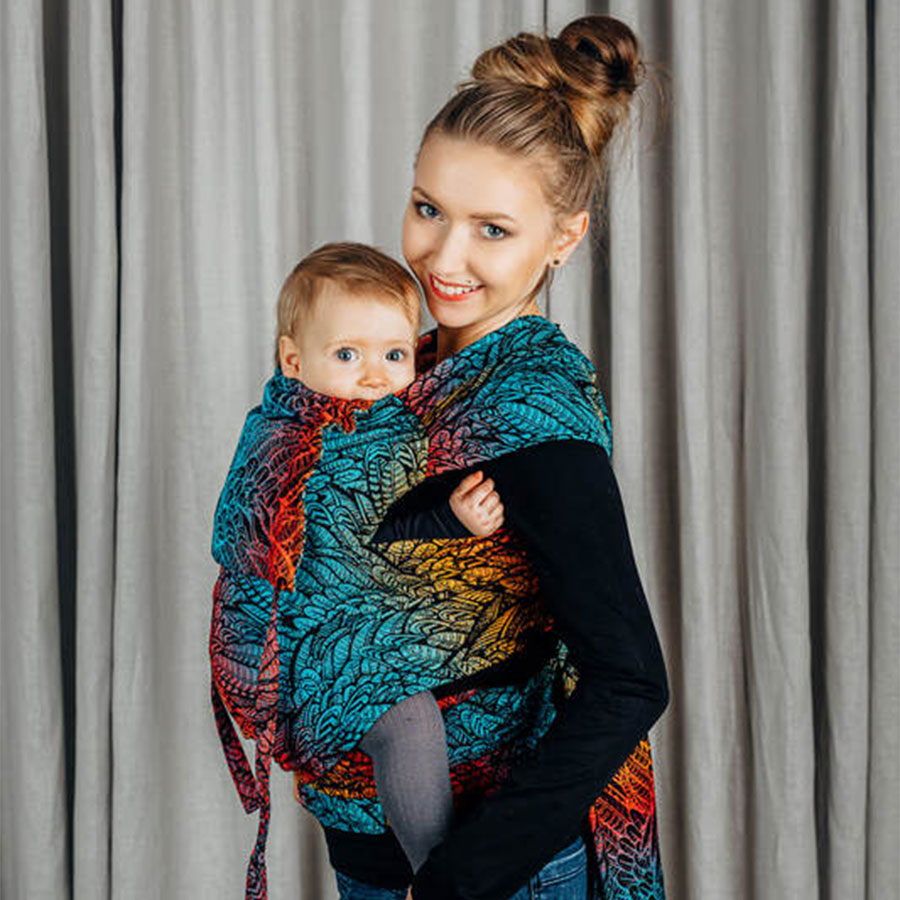 A woman with brown hair standing indoors carrying her toddler on her front in a LennyLamb Wraptai