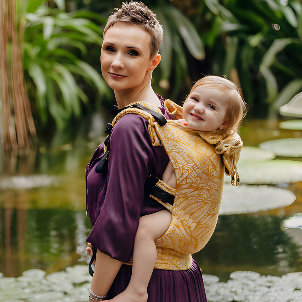 A woman with short hair wearing a purple dress, standing beside a pond carrying her smiling baby girl on her back in a Lenny Lamb Go Carrier