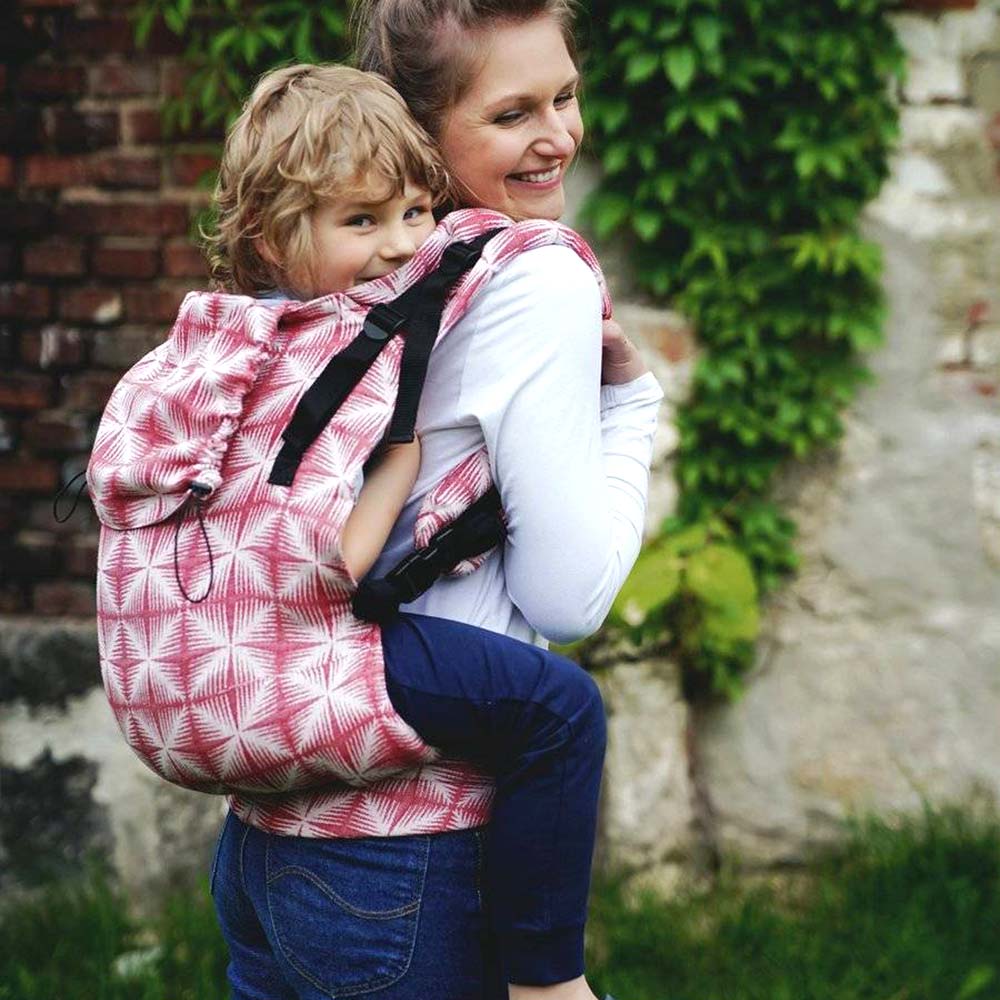 A woman with brown hair wearing a white top and blue jeans, standing on a path in front of a red brick and white painted wall, looking over her right shoulder smiling at the camera, carrying her son on her back in a Little Frog Toddler XL Carrier Red Glimmer 