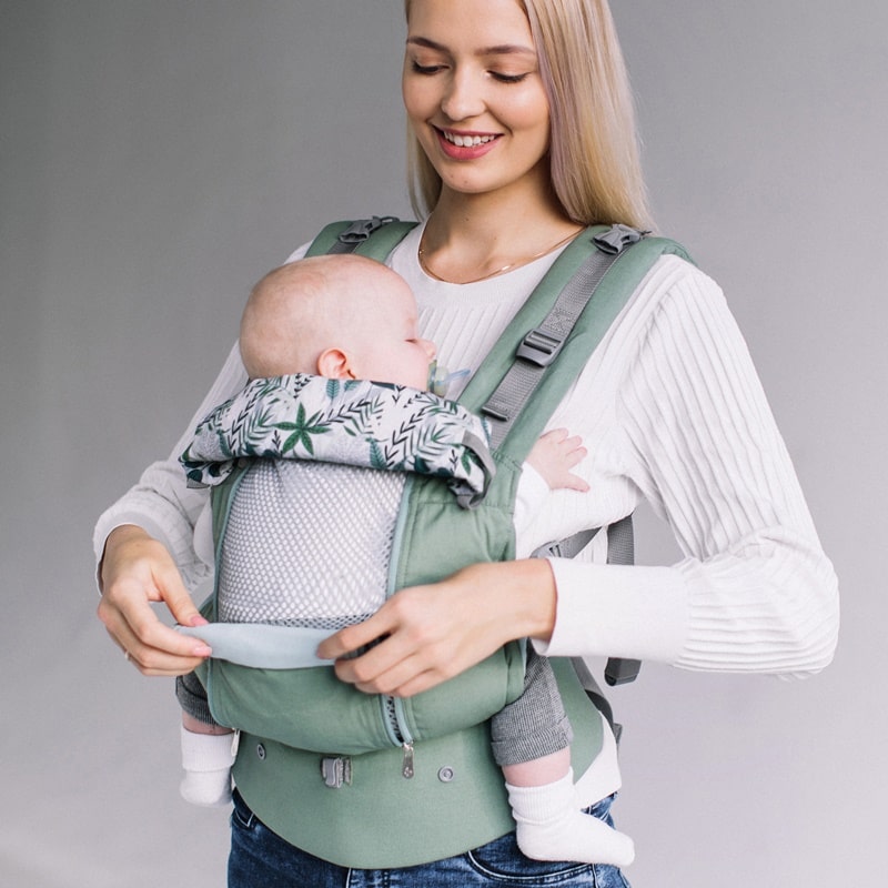 A woman with blond hair wearing a white top and blue jeans, demonstrating the rollaway panel of the Love and Carry AIR X baby carrier, whilst her baby naps quietly in the carrier.