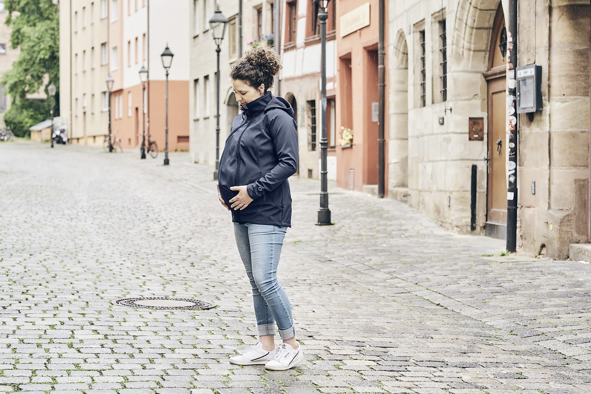 A woman with brown curly hair done up in a bun standing in the middle of a cobbled street looking down lovingly at her pregnancy bump whilst wearing the Mamalila Babywearing jacket with the maternity insert added.