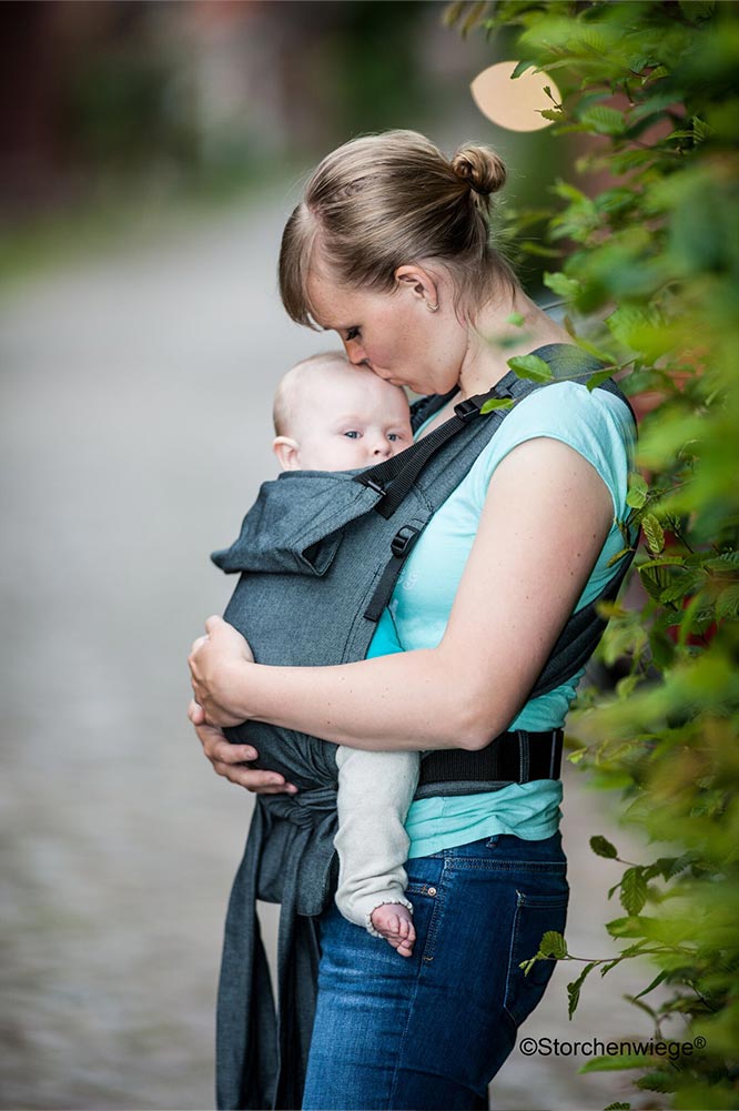 A woman wearing a white t-shirt and blue jeans standing on a path beside a hedge kissing the top of her baby's head whilst she carries them in a Storchenwiege baby carrier 